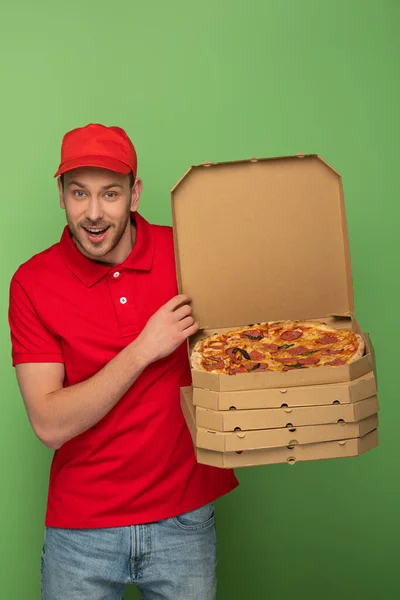 Взволнованный курьер в красной форме держит коробки пиццы на зеленом — стоковое фото
