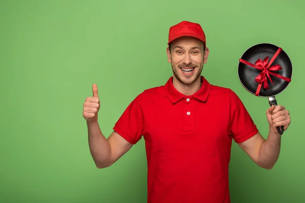 Felice uomo di consegna in uniforme rossa tenendo padella con fiocco e mostrando pollice in su sul verde — Foto stock