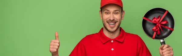 Happy delivery man in red uniform holding frying pan with bow and showing thumb up isolated on green — Stock Photo