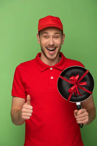 Happy delivery man in red uniform holding frying pan with bow and showing thumb up on green — Stock Photo