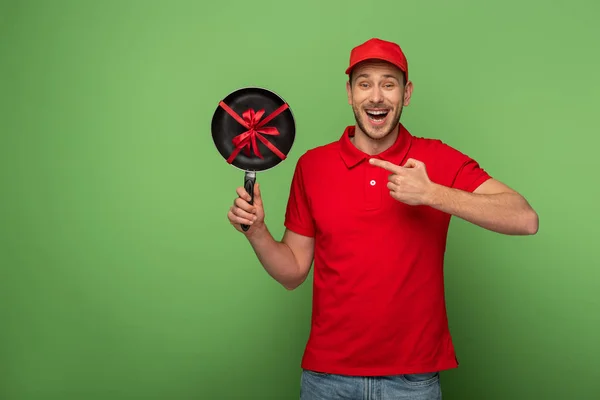 Щасливий чоловік доставки в червоній формі вказуючи пальцем на сковороді з бантом на зеленому — стокове фото
