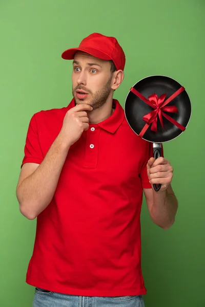Pensive delivery man in red uniform holding frying pan with bow on green — Stock Photo