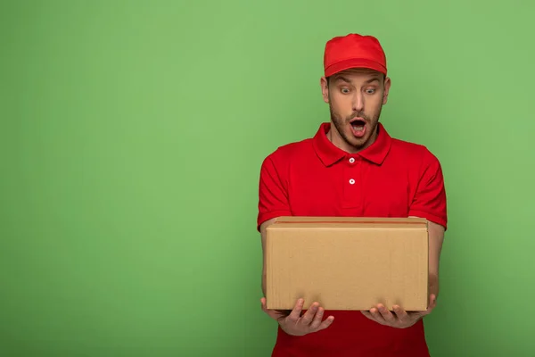 Шокований чоловік доставки в червоній формі тримає посилку на зеленому — стокове фото