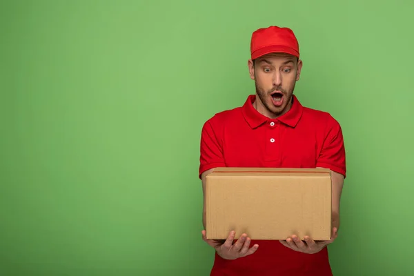 Sorprendido repartidor en rojo uniforme holding paquete en verde - foto de stock