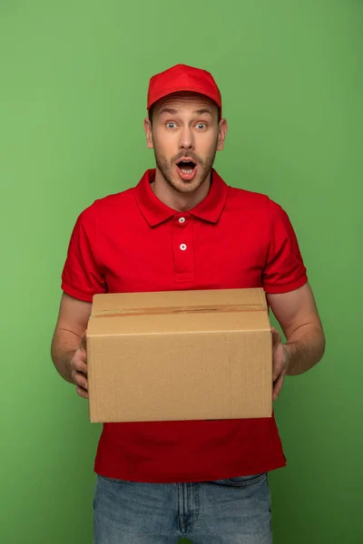 Sorprendido repartidor en rojo uniforme holding paquete en verde — Stock Photo