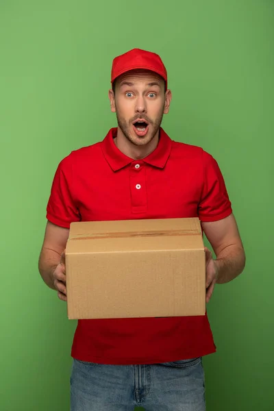 Sorprendido repartidor en rojo uniforme holding paquete en verde - foto de stock