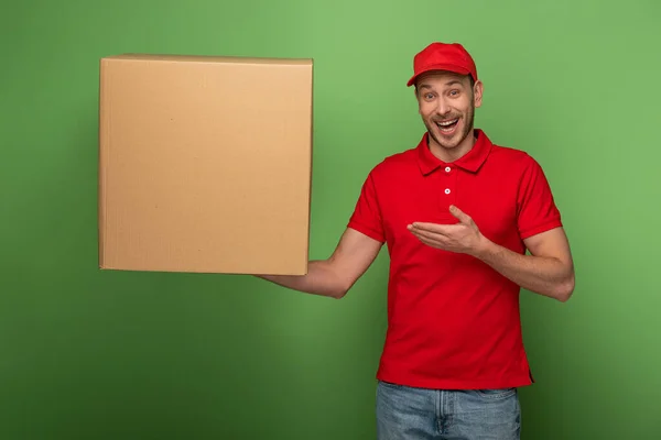 Glücklicher Zusteller in roter Uniform zeigt mit der Hand auf riesiges Paket auf Grün — Stockfoto