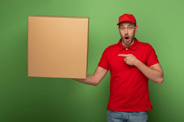 Шокований чоловік доставки в червоній формі вказує пальцем на величезну посилку на зеленому — стокове фото