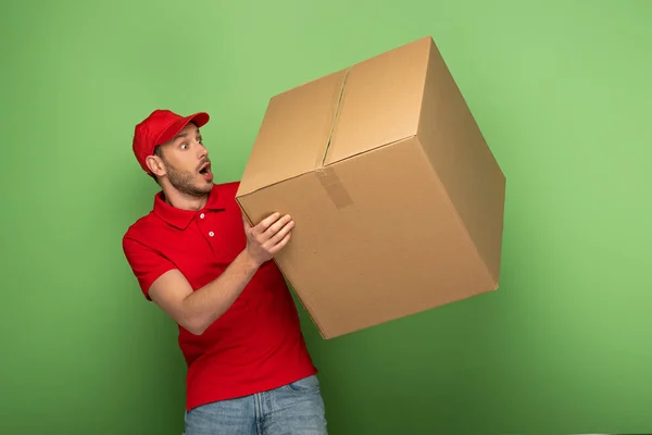 Chocado repartidor hombre en rojo uniforme celebración enorme paquete en verde - foto de stock