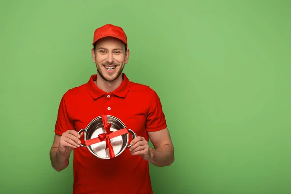 Happy delivery man in red uniform holding pan with bow on green — Stock Photo