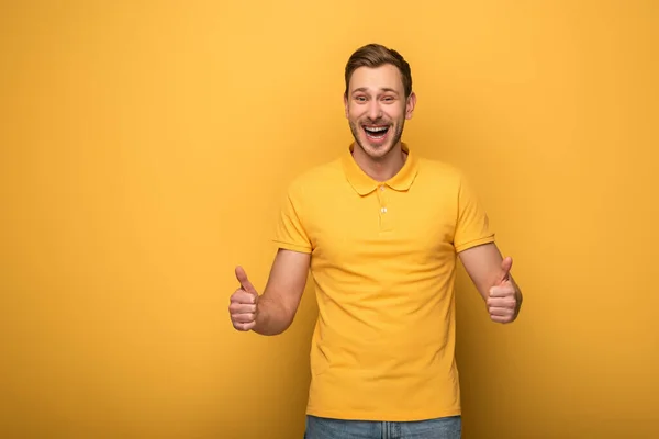 Homem bonito feliz em roupa amarela mostrando polegares para cima no fundo amarelo — Fotografia de Stock