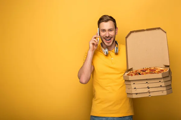 Happy man with headphones in yellow outfit holding pizza and talking on smartphone on yellow background — Stock Photo