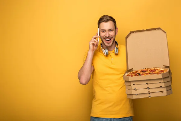 Glücklicher Mann mit Kopfhörern in gelbem Outfit, der Pizza hält und auf gelbem Hintergrund mit dem Smartphone spricht — Stockfoto