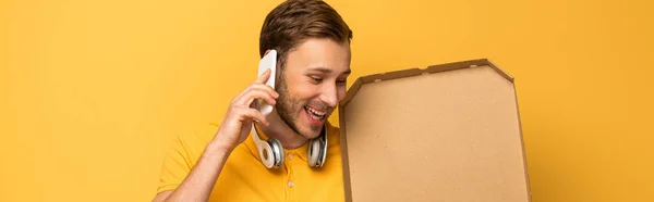 Happy man with headphones in yellow outfit holding pizza box and talking on smartphone isolated on yellow, panoramic shot — Stock Photo