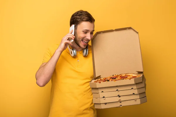 Happy man with headphones in yellow outfit holding pizza and talking on smartphone on yellow background — Stock Photo