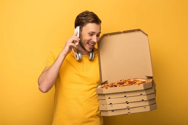 Homem feliz com fones de ouvido em roupa amarela segurando pizza e falando no smartphone no fundo amarelo — Fotografia de Stock