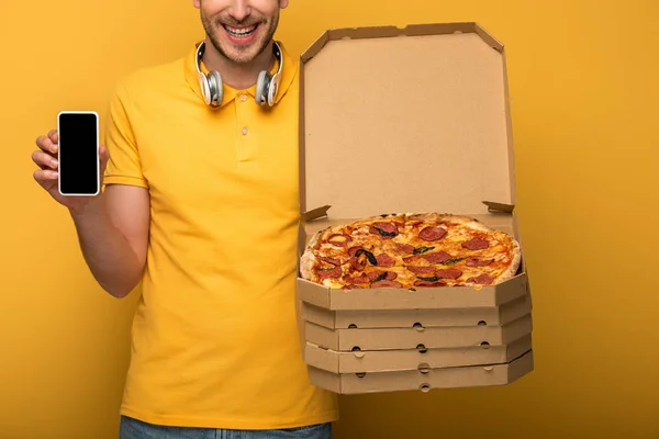 Cropped view of happy man with headphones in yellow outfit holding pizza and smartphone on yellow background — Stock Photo