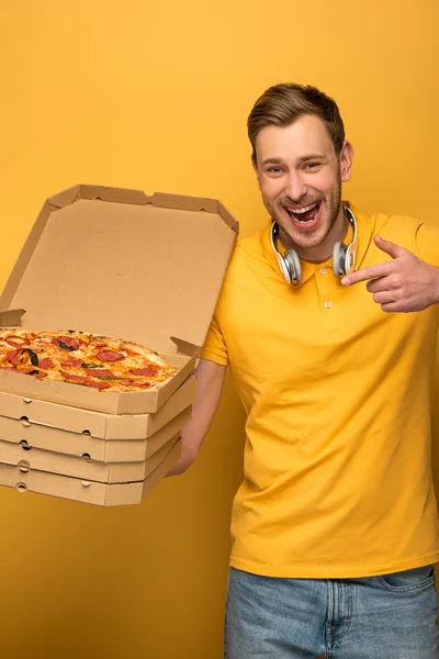 Hombre feliz con auriculares en traje amarillo apuntando a la pizza sobre fondo amarillo - foto de stock