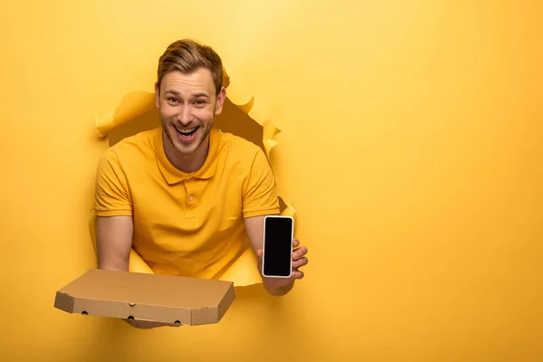 Happy handsome man in yellow outfit holding smartphone and pizza box in yellow paper hole — Stock Photo