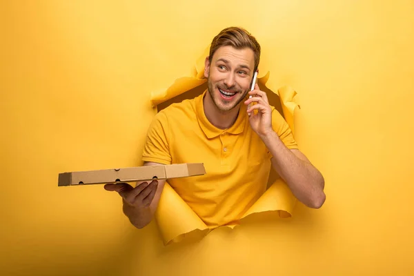 Hombre guapo feliz en traje amarillo hablando en el teléfono inteligente y sosteniendo la caja de pizza en agujero de papel amarillo - foto de stock