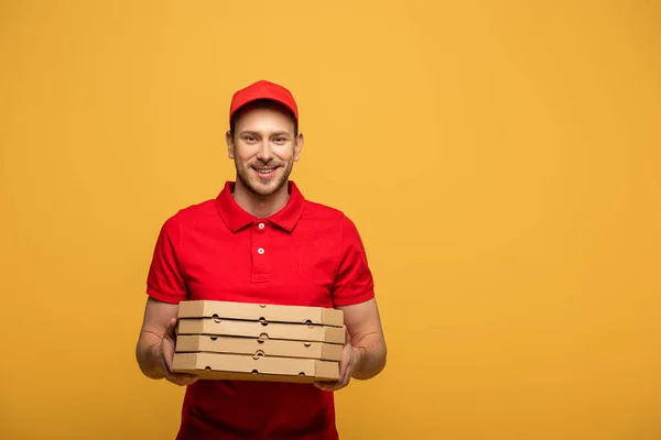 Homem de entrega feliz em uniforme vermelho segurando caixas de pizza isoladas no amarelo — Fotografia de Stock