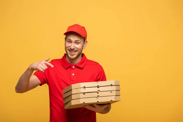 Homem de entrega feliz em uniforme vermelho apontando com o dedo para caixas de pizza isoladas em amarelo — Fotografia de Stock