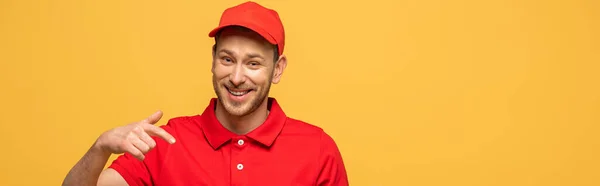 Feliz repartidor en uniforme rojo apuntando con el dedo aislado en amarillo, tiro panorámico - foto de stock