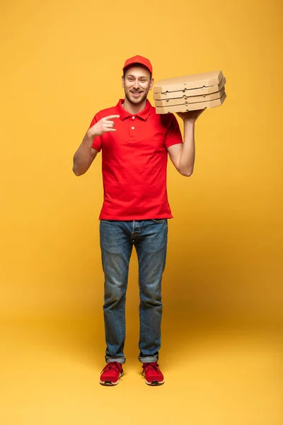 Happy delivery man in red uniform pointing with finger at pizza boxes on yellow — Stock Photo