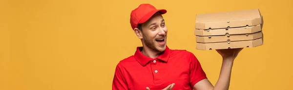 Happy delivery man in red uniform pointing with hand at pizza boxes isolated on yellow, panoramic shot — Stock Photo