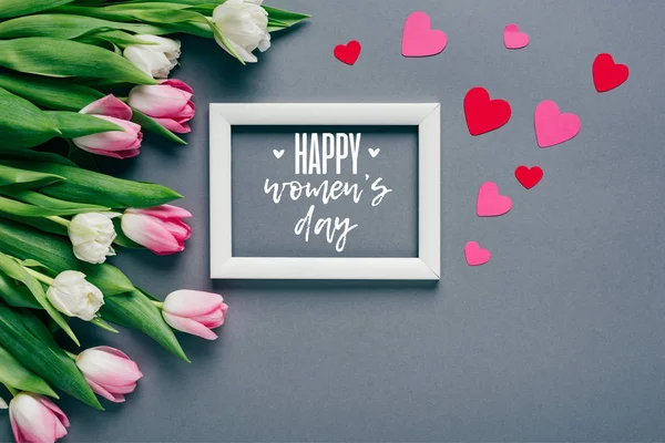 Top view of white frame with happy womens day lettering near paper hearts and tulips on grey background — Stock Photo