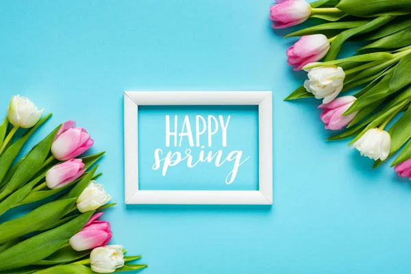 Draufsicht auf weißen Rahmen mit fröhlichem Frühlingsschriftzug in der Nähe von Tulpen auf blauem Hintergrund — Stockfoto