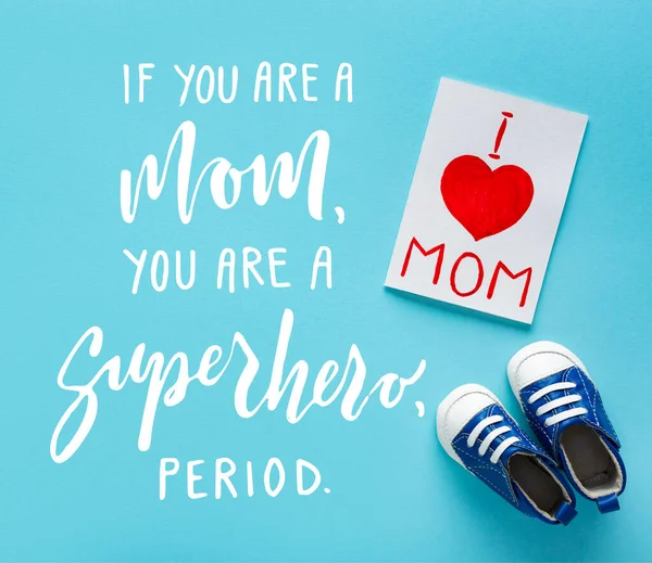 Vista superior de la tarjeta de felicitación con letras de mamá me encanta cerca de botines de bebé y si usted es mamá, usted es un superhéroe, período de letras sobre fondo azul - foto de stock