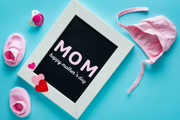 Kreidetafel mit Mama-Schriftzug in der Nähe von rosa Babykleidung und Schnuller auf blauem Hintergrund — Stockfoto