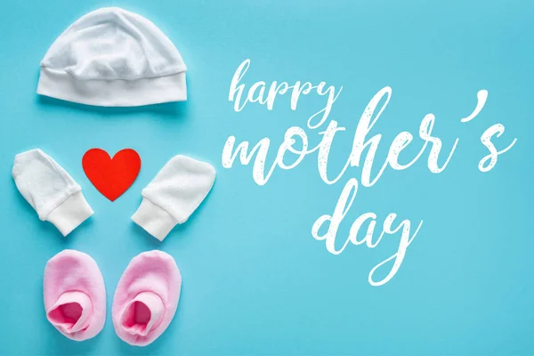 Vista superior de manoplas de bebé, botines y sombrero con corazón de papel sobre fondo azul, ilustración feliz día de las madres - foto de stock