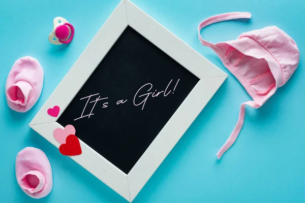 Top view of empty chalkboard with it is a girl lettering near pink baby clothes and pacifier on blue background — Stock Photo