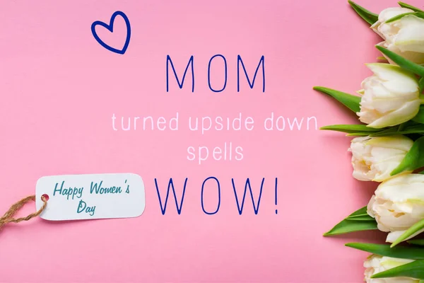 Top view of paper label with happy womens day lettering near white tulips on pink background, mom wow illustration — Stock Photo