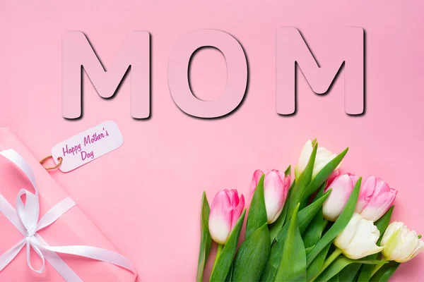 Top view of gift with happy mothers day lettering on paper label near tulips on pink background, mom illustration — Stock Photo