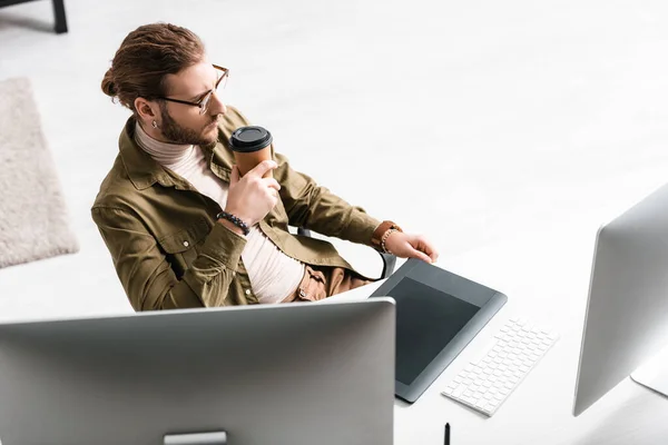 Высокоугольный вид цифрового дизайнера, пьющего кофе рядом с компьютерами и графическим столиком на столе — стоковое фото