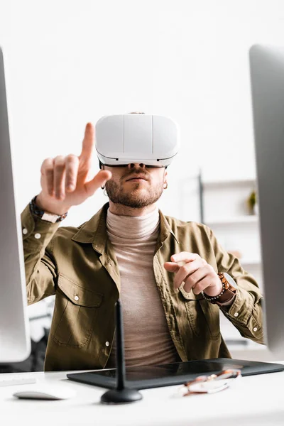 Foco seletivo do designer digital em fones de ouvido de realidade virtual apontando com o dedo perto de gráficos tablet e computadores na mesa — Fotografia de Stock