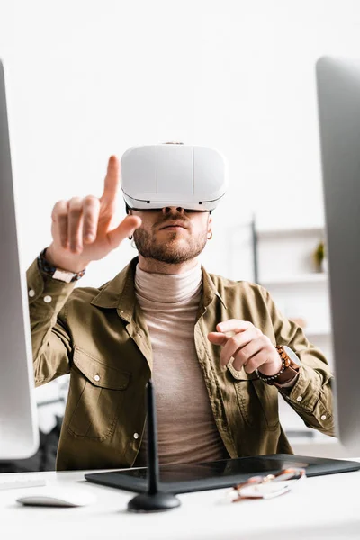 Selective focus of digital designer in virtual reality headset pointing with finger near graphics tablet and computers on table — Stock Photo