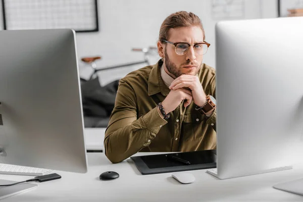 Pensativo artista 3d mirando a la cámara cerca de computadoras y gráficos tableta en la mesa en la oficina - foto de stock