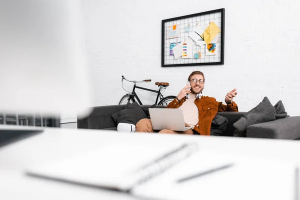 Foco seletivo de sorrir artista 3d falando no smartphone e segurando laptop no sofá no escritório — Fotografia de Stock