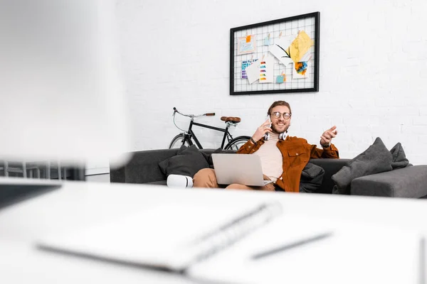 Focus selettivo di artista 3D sorridente che parla su smartphone e tiene il computer portatile sul divano in ufficio — Foto stock