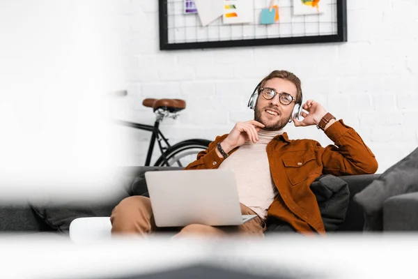 Selective focus of smiling digital designer in headphones holding laptop near vr headset on couch in office — Stock Photo