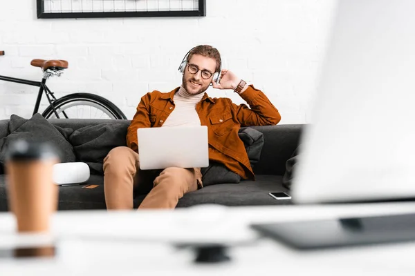 Selective focus of smiling digital designer in headphones using laptop near vr headset and smartphone on couch — Stock Photo