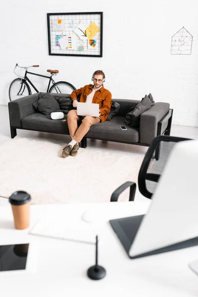Selective focus of 3d artist in headphones using laptop near smartphone and virtual reality headset on couch in office — Stock Photo