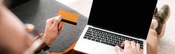 Selective focus of digital designer using laptop and holding credit card on couch, panoramic shot — Stock Photo