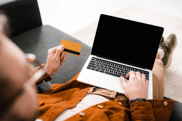 Foco seletivo do designer digital usando laptop com tela em branco e segurando cartão de crédito no sofá — Fotografia de Stock