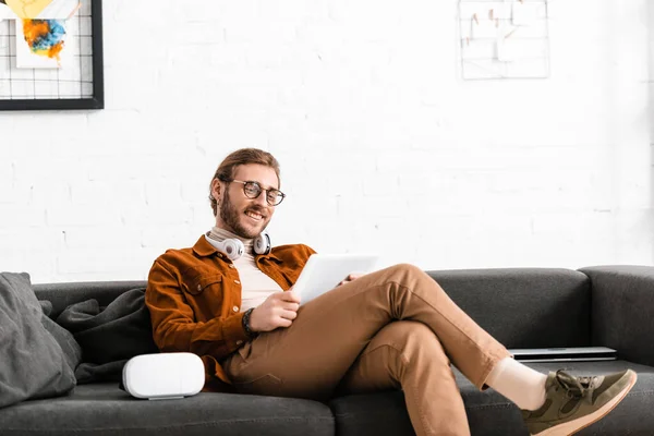 Lächelnder digitaler Designer mit digitalem Tablet in der Nähe von vr Headset und Laptop auf der Couch — Stockfoto