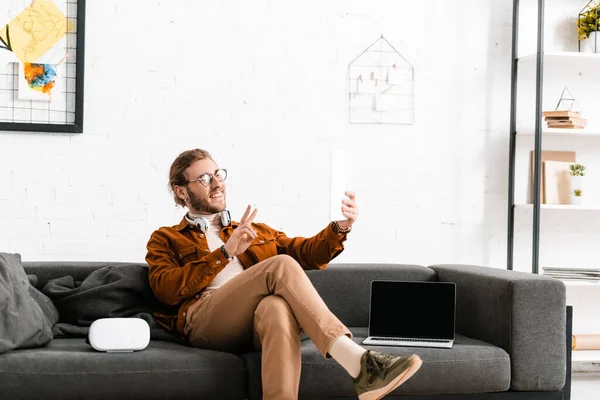 Sorrindo 3d artista mostrando símbolo de paz ao ter chamada de vídeo em tablet digital no sofá no escritório — Fotografia de Stock